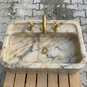 May include: Rectangular marble sink with a gold tap and two gold cross-shaped handles. The sink has a cream and brown marbled pattern. The drain is gold. The sink is on a wooden surface.