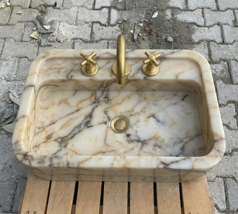 May include: Rectangular marble sink with a gold tap and two gold cross-shaped handles. The sink has a cream and brown marbled pattern. The drain is gold. The sink is on a wooden surface.