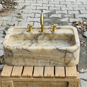 May include: A rectangular marble sink with a gold tap and two gold handles. The sink has a beige and brown veined pattern. It is sitting on a wooden pallet.