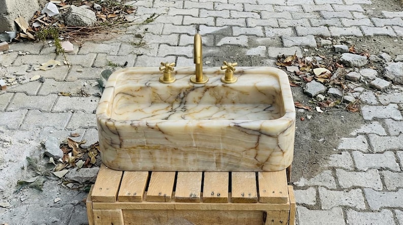 May include: A rectangular marble sink with a gold tap and two gold handles. The sink has a beige and brown veined pattern. It is sitting on a wooden pallet.