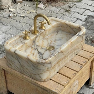 May include: A rectangular marble sink with a gold tap and fixtures. The sink has a light beige and brown veined pattern. The sink is sitting on a wooden pallet.