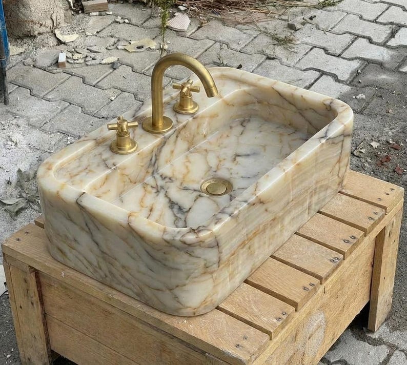 May include: A rectangular marble sink with a gold tap and fixtures. The sink has a light beige and brown veined pattern. The sink is sitting on a wooden pallet.