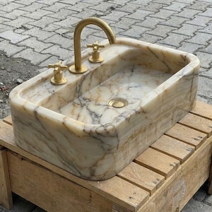 May include: A rectangular marble sink with a gold tap and fixtures. The sink features a natural stone pattern with beige and grey veining. The tap is curved, and the fixtures are cross-shaped. The sink is set on a wooden stand.
