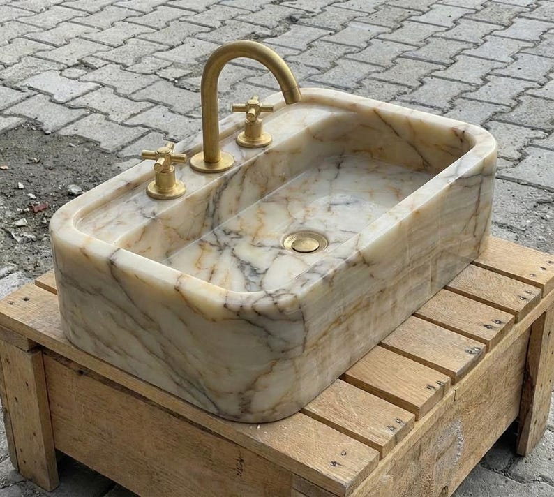 May include: A rectangular marble sink with a gold tap and fixtures. The sink features a natural stone pattern with beige and grey veining. The tap is curved, and the fixtures are cross-shaped. The sink is set on a wooden stand.