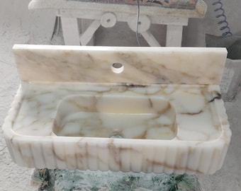 Puede incluir: Un lavabo de baño de mármol blanco con un solo agujero para un grifo. El lavabo tiene un borde decorativo acanalado y una encimera de mármol blanco.