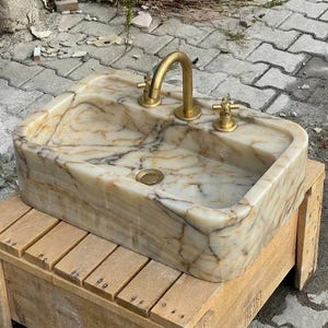 May include: A rectangular marble sink with a gold tap and fixtures. The sink has a beige and brown marbled pattern. The tap is a curved gooseneck style. The sink is sitting on a wooden pallet.