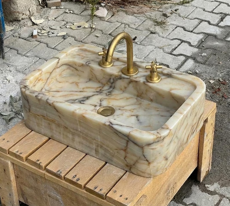 May include: A rectangular marble sink with a gold tap and fixtures. The sink has a beige and brown marbled pattern. The tap is a curved gooseneck style. The sink is sitting on a wooden pallet.