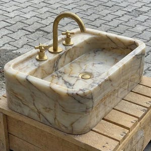 May include: A rectangular marble sink with a gold tap and two gold handles. The sink has a natural stone pattern with beige, brown, and white veining. The sink is on a wooden crate.
