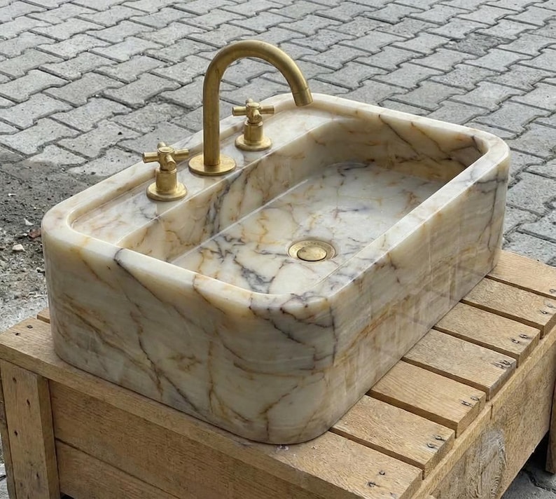 May include: A rectangular marble sink with a gold tap and two gold handles. The sink has a natural stone pattern with beige, brown, and white veining. The sink is on a wooden crate.