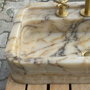 May include: A rectangular marble sink with a cream and brown veined pattern. The sink has rounded corners and is fitted with gold-coloured taps and a drain. The overall design is luxurious and elegant.