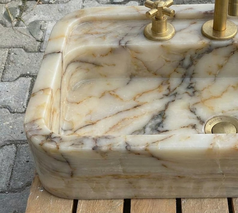 May include: A rectangular marble sink with a cream and brown veined pattern. The sink has rounded corners and is fitted with gold-coloured taps and a drain. The overall design is luxurious and elegant.