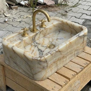 May include: A rectangular onyx stone sink with a gold-coloured tap and fixtures. The sink has a natural, marbled pattern in cream, beige, and brown. The sink is mounted on a wooden stand.