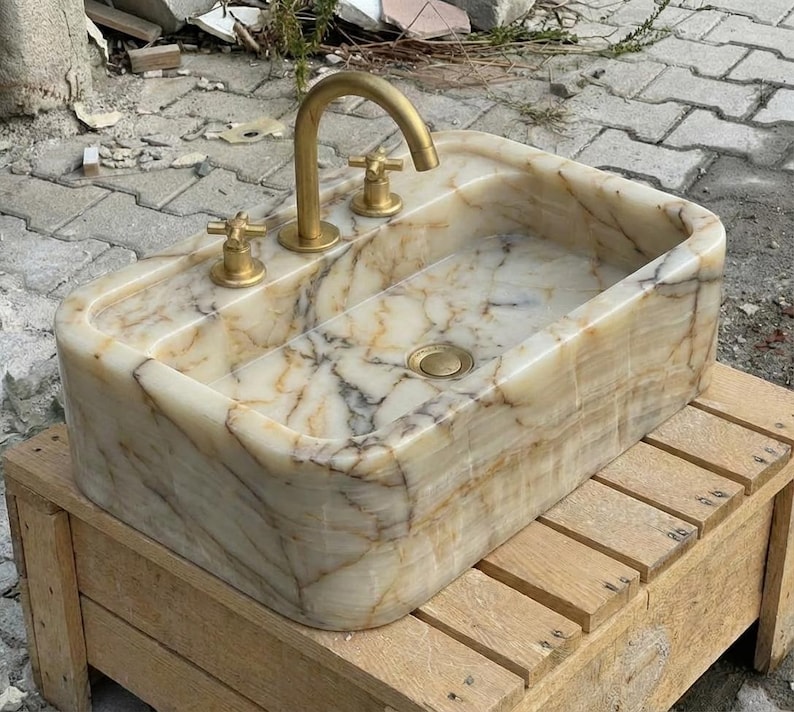 May include: A rectangular onyx stone sink with a gold-coloured tap and fixtures. The sink has a natural, marbled pattern in cream, beige, and brown. The sink is mounted on a wooden stand.