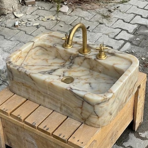 May include: Rectangular marble sink with a gold tap and fixtures. The sink has a beige and brown veined pattern. The tap is a gooseneck style. The sink is sitting on a wooden crate.