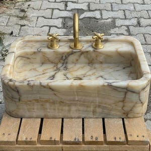 May include: Rectangular marble sink with gold-coloured tap and handles. The sink has a beige and brown veined pattern. The sink is sitting on a wooden pallet.