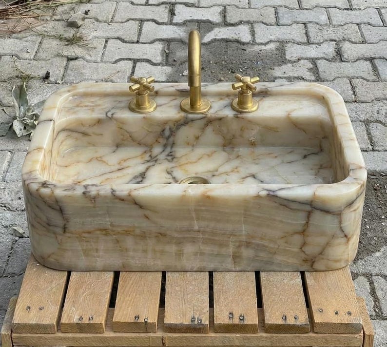 May include: Rectangular marble sink with gold-coloured tap and handles. The sink has a beige and brown veined pattern. The sink is sitting on a wooden pallet.