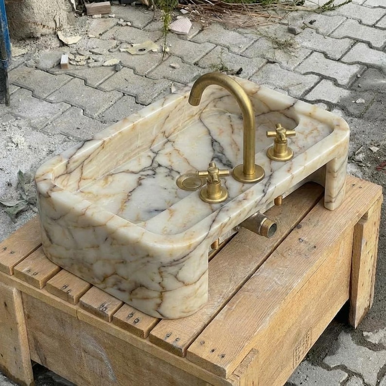 May include: A rectangular marble sink with a gold tap and fixtures. The sink has a cream and brown veined pattern. The tap is curved, and the fixtures include two cross-shaped handles. The sink is set on a wooden crate.