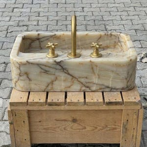 May include: A rectangular onyx stone sink with a gold tap and two gold cross-handle taps. The sink has a light beige and brown marbled pattern. The sink is on a wooden pallet.