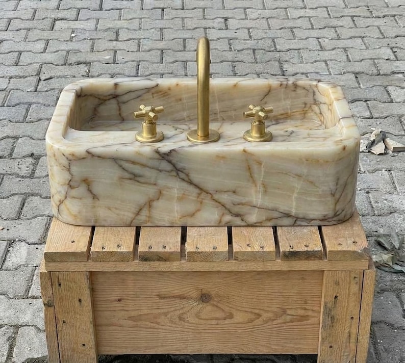 May include: A rectangular onyx stone sink with a gold tap and two gold cross-handle taps. The sink has a light beige and brown marbled pattern. The sink is on a wooden pallet.