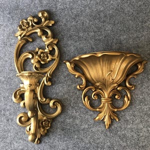 Puede incluir: Dos apliques de pared dorados y ornamentados. Uno presenta un candelabro y detalles florales, mientras que el otro tiene forma de cuenco decorativo. Ambos tienen un estilo barroco vintage con intrincados adornos y un acabado metálico.