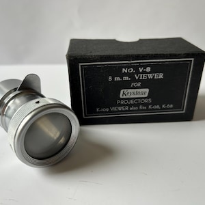 May include: A vintage Keystone 8mm film viewer with a silver metal body and a black box. The box has white text that reads "NO. V-8 8 m.m. VIEWER FOR Keystone PROJECTORS". The viewer is designed for K-109, K-08, and K-68 projectors.