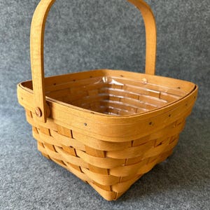 Può includere: Un cestino quadrato marrone chiaro con un manico curvo. Il cestino è realizzato con strisce di legno intrecciate e presenta un rivestimento in plastica trasparente. Il manico è fissato con piccoli pioli di legno. Il cestino è vuoto.
