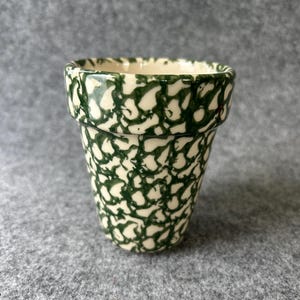 May include: A small, ceramic flower pot with a green and white pattern. The pot has a slightly flared rim and a cylindrical body. The green pattern is a repeating, abstract design. The pot is empty and sits on a gray surface.