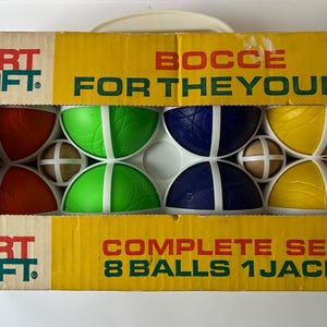 以下が含まれることがあります： 黄色とベージュの段ボール箱に入った、ヴィンテージのSportcraftボッチェボールセット。「Bocce for the Young」と「Complete Set 8 Balls 1 Jack」と書かれています。セットには、赤、緑、青、黄色のボールと、小さな白と茶色のジャックが含まれています。