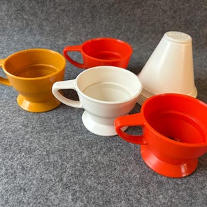 Puede incluir: Un surtido de tazas de plástico de colores y un embudo blanco. Las tazas son en tonos de naranja, amarillo y blanco, cada una con un asa y una base de pedestal. El embudo blanco tiene forma de cono.