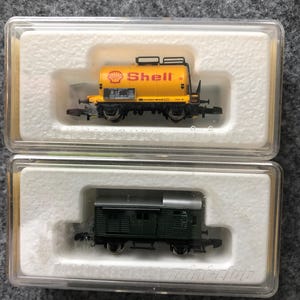 May include: Two model train cars in clear plastic cases. One is a yellow Shell tanker car, and the other is a dark green boxcar. Both are miniature and detailed, with small wheels and intricate designs.