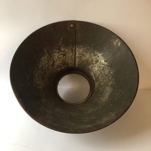 Puede incluir: Un embudo metálico circular y envejecido, con un borde ancho y acampanado y una abertura central. El metal presenta un aspecto oscuro y desgastado, con desgaste visible y un pequeño agujero cerca del borde superior.