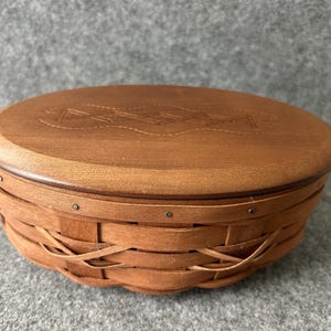 May include: An oval-shaped wooden basket with a smooth, solid wood lid featuring a decorative etched pattern. The base is constructed from woven wooden strips secured with small metal nails along the upper rim, creating a sturdy container.