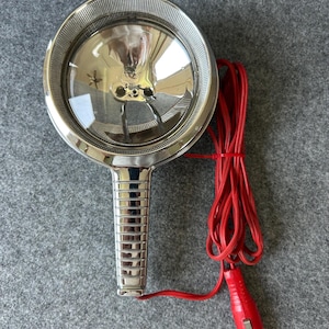 May include: A handheld chrome spotlight featuring a large circular reflector lens and a ribbed handle. The device is connected to a long red power cord that terminates in a red 12-volt automotive accessory plug, designed for use with a vehicle power outlet.