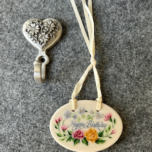 May include: A heart-shaped metal hook with floral detailing and a small oval ceramic ornament. The ornament has a floral design with the words "Happy Birthday" in script. The ornament is cream-coloured with a ribbon.