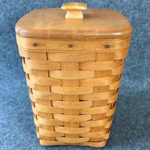 May include: A square woven basket with a light brown finish and a matching wooden lid. The lid features a small, centered square knob. The basket body is constructed from flat, interlaced strips of wood, secured at the top rim with small metal nails.