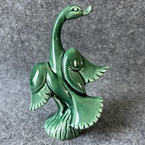 May include: A glossy, dark green ceramic figurine of a goose with its wings spread wide. The bird is depicted in a dynamic pose with an elongated neck and a textured base, featuring a smooth, reflective glaze that highlights its curved form.
