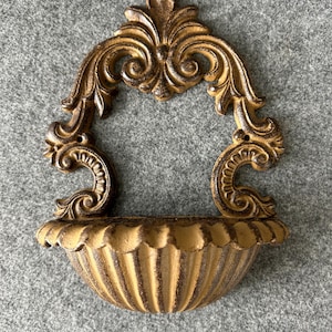 May include: Ornate, antique-style wall sconce in a warm gold finish. The sconce features a decorative, arched top with scrolling details and a ribbed, bowl-shaped base, perfect for displaying small items or plants.