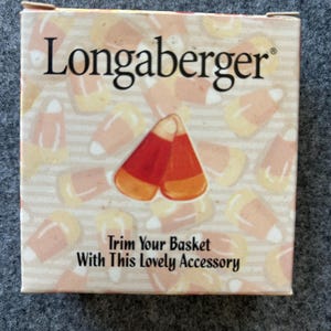 Può includere: Una scatola quadrata di cartone con il nome del marchio Longaberger in nero. Lo sfondo presenta un motivo ripetuto di caramelle a forma di mais arancioni, gialle e bianche. Il testo recita, Trim Your Basket With This Lovely Accessory.