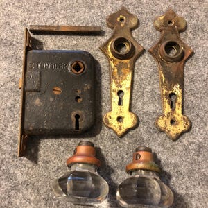 May include: An assortment of antique door hardware, including a black metal lock with the text "S. TUMBLER", two ornate brass backplates, two clear glass doorknobs with copper-colored tops, and a metal strike plate.