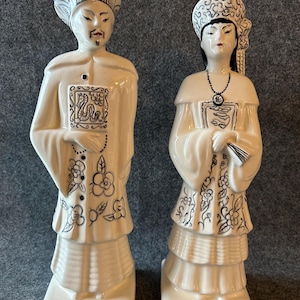Puede incluir: Dos figuras de cerámica de personajes asiáticos con vestimenta tradicional. Las figuras son de color blanquecino con detalles en negro, incluyendo patrones florales y rasgos faciales. La figura masculina sostiene un libro, y la femenina un abanico.