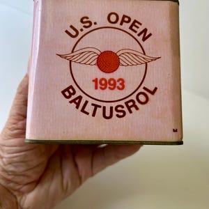 以下が含まれることがあります： ピンク色の金属製の箱で、茶色の文字で「U.S. OPEN BALTUSROL 1993」と書かれています。箱には翼のあるゴルフボールのロゴが付いています。箱には金色のトリムが付いています。
