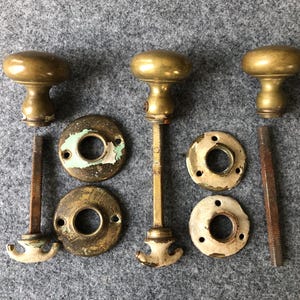 May include: Three antique brass door knobs with matching hardware. The knobs are a warm gold color, and the hardware includes round backplates and threaded rods. The pieces show signs of age and wear, with some paint chipping.
