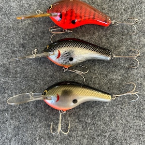 May include: Three colorful fishing lures. The top lure is red with black accents, the middle one is silver and black, and the bottom one is gold and silver. Each lure has a clear plastic lip and multiple treble hooks.