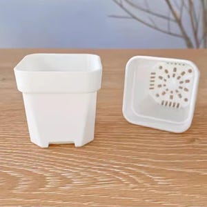 May include: A white square-shaped plant pot with a matching drainage tray. The pot has a geometric design and is sitting on a wooden surface. The tray has a series of holes for drainage.
