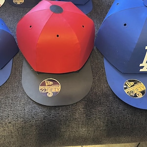 Könnte beinhalten: Drei Baseballkappen in Blau, Rot und Grau. Die blaue Kappe links hat ein Buffalo Bills-Logo, die rote Kappe ist schlicht und die blaue Kappe rechts hat ein weißes "LA"-Logo. Alle haben einen 59FIFTY-Aufkleber.