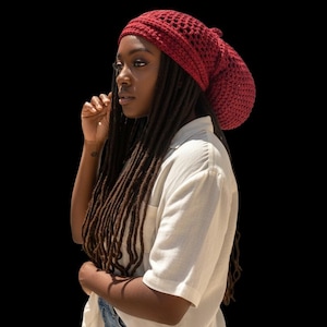 May include: A burgundy crochet beanie with a textured design. The hat is worn with a white button-down shirt and denim shorts. The background is solid black.