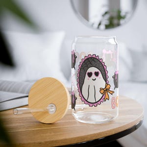 May include: A clear glass tumbler with a bamboo lid and a glass straw. The glass features a graphic of a white ghost wearing pink heart-shaped sunglasses inside a black oval frame with a pink scalloped border and an orange bow, plus black cauldrons.