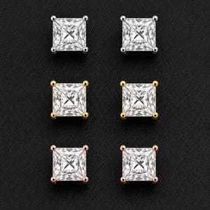 May include: Three pairs of square-cut stud earrings arranged in a vertical column. The top pair is silver-toned, the middle pair is yellow gold-toned, and the bottom pair is rose gold-toned. Each earring features a clear, faceted stone with four prongs.