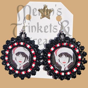 May include: A pair of handmade earrings featuring a circular design with a portrait of a woman wearing a Santa hat. The earrings have a black beaded border with red and white accents, and silver-tone ear wires.
