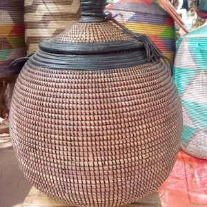 May include: A large, round, woven basket with a black lid and a black tassel. The basket is made of natural materials and has a brown and black pattern.